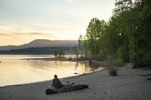 Una persona sentada sobre un tronco junto a un lago, con árboles y una montaña al fondo.