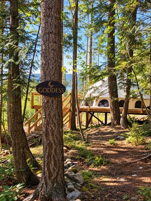 A forested area with a sign reading 'GODDESS' and a wooden structure with a view of a tent-like building. - Goddess at Namastay Glamping Retreat (Columbia-Shuswap)