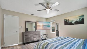 Quarto com cama, penteadeira, ventilador de teto e quadros nas paredes.