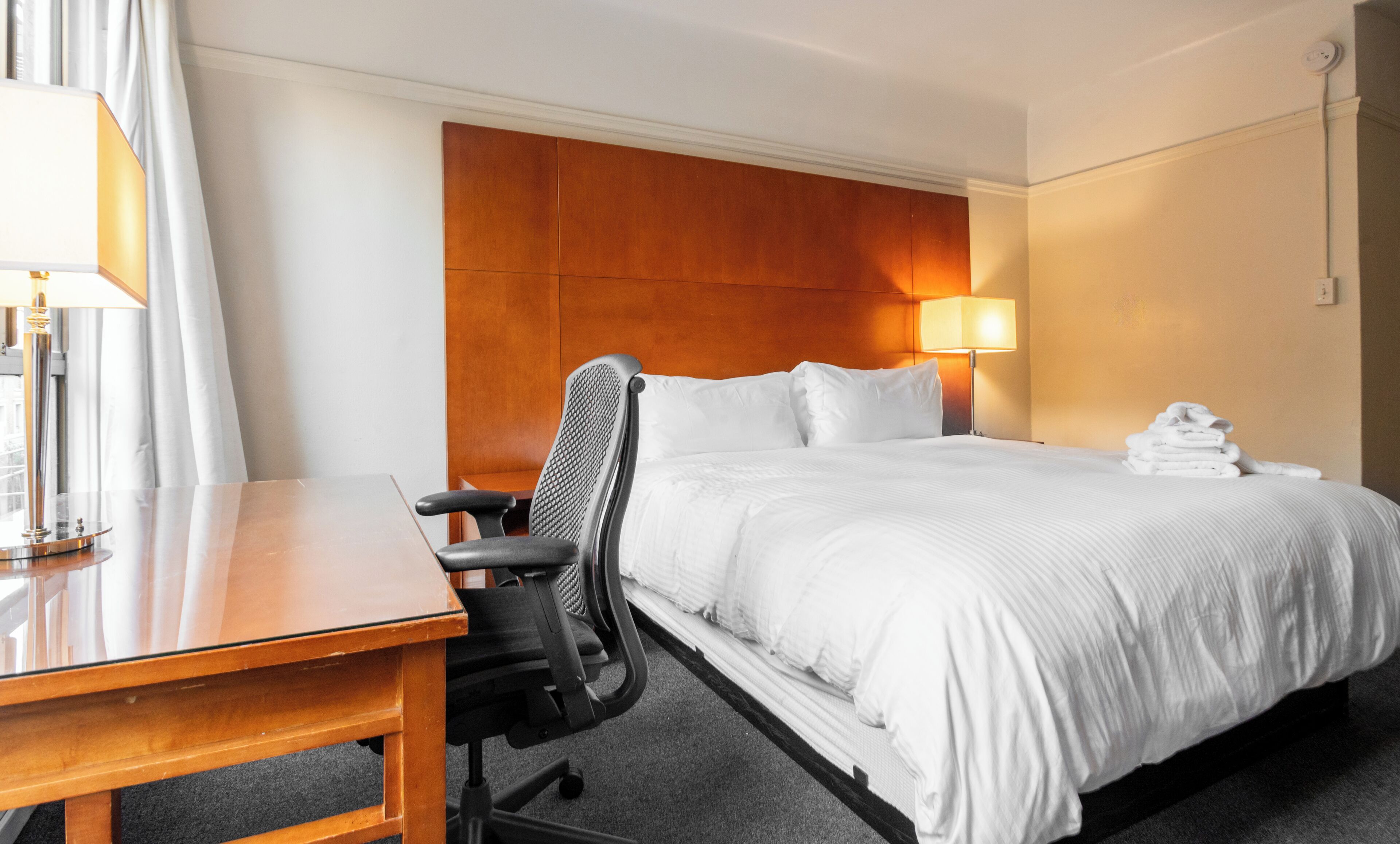 Une chambre d’hôtel avec un lit, un bureau, une chaise, une lampe et une pile de serviettes.
