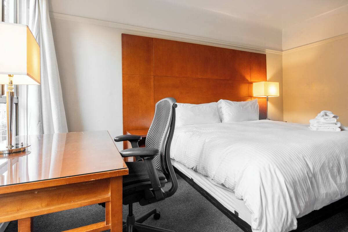 A hotel room with a bed, a desk, a chair, a lamp, and a stack of towels.