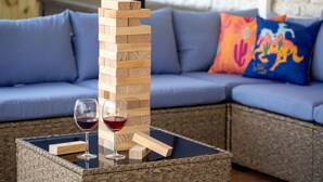 Uma torre de Jenga de madeira sobre uma mesa de vidro, com dois copos de vinho tinto, colocada sobre uma mesa de vime em frente a um sofá azul com almofadas coloridas.