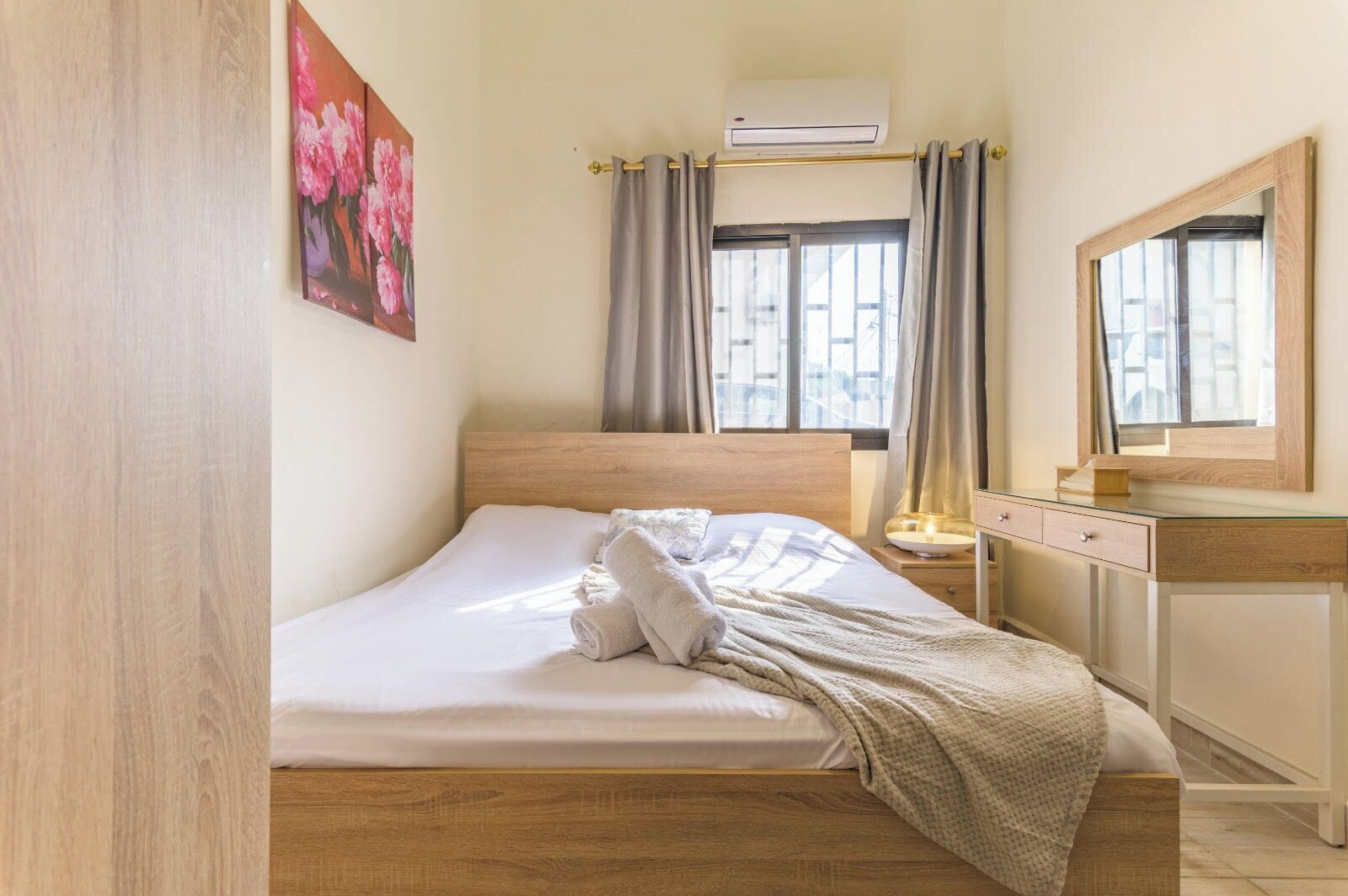 Quarto de hotel com cama de madeira, mesa de cabeceira com luminária, espelho e janela com cortinas.
