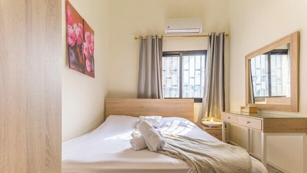 A hotel room with a wooden bed, a bedside table with a lamp, a mirror, and a window with curtains. - Daya 2BR Apartment In Batroun (Batroun)
