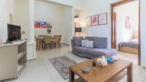 A modern living room with a grey sofa, a coffee table, and a TV on a stand. There is a dining area with a table and chairs, and a small rug on the floor. - Daya 2BR Apartment In Batroun (Batroun)