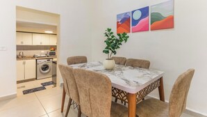 A dining area with a marble table, beige chairs, and a potted plant. A kitchen is visible in the background with a washing machine and a microwave. - Daya 2BR Apartment In Batroun (Batroun)