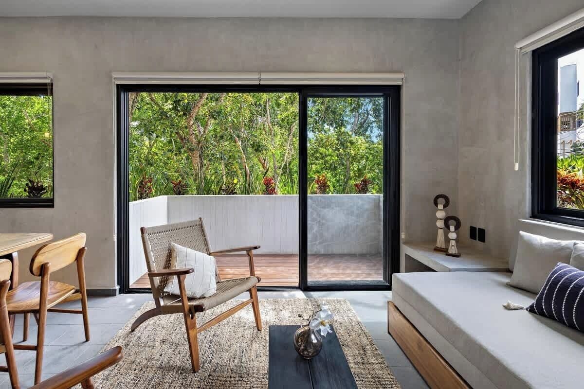 A modern living room with a sofa, wooden chairs, a small table, and a large sliding glass door leading to a balcony with a view of greenery.