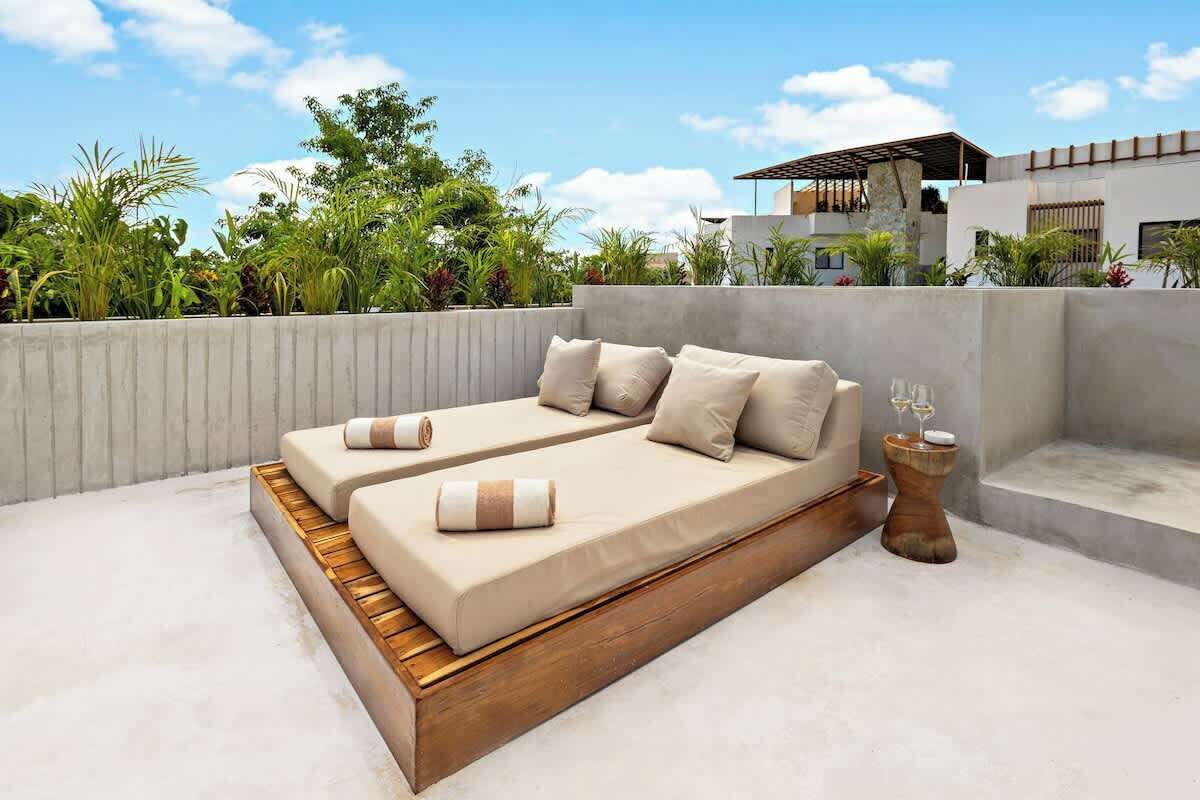 Ein moderner Sitzbereich im Freien mit einer Couch und Kissen, einem Holztisch mit einem Glas sowie Blick auf ein Gebäude und Grünflächen.