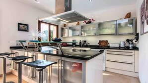 A modern kitchen with a central island, bar stools, stainless steel appliances, and a large window. - Holiday Home 'Amarador' with Private Pool, Wi-Fi and Air Conditioning (Santanyí)