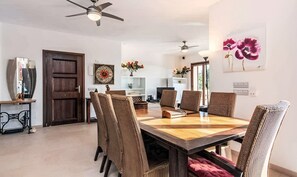 A dining area with a wooden table and woven chairs, a ceiling fan, and a painting of flowers on the wall.