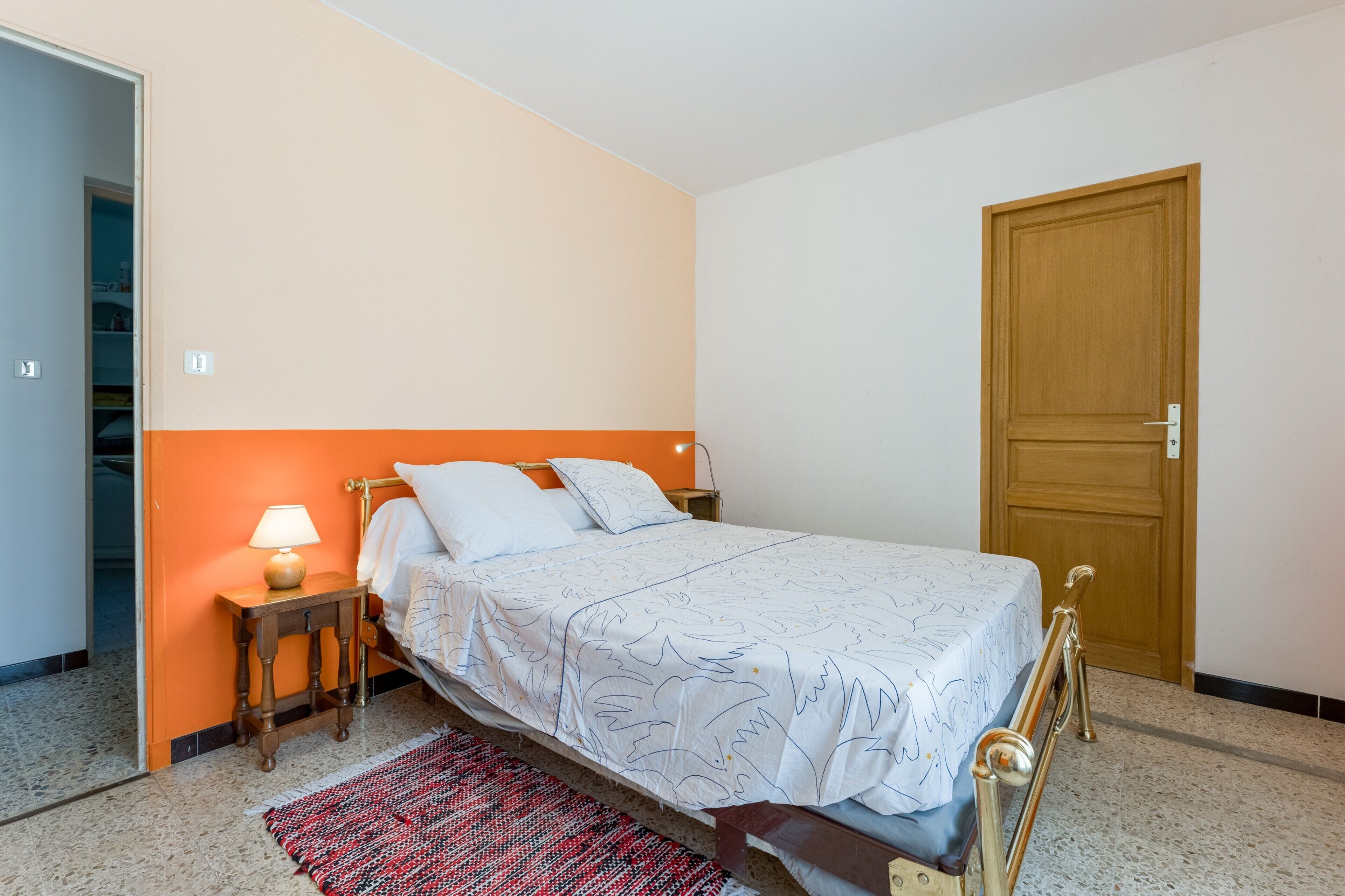 Un dormitorio con una cama, una mesita de noche, una lámpara, una puerta y una alfombra estampada.