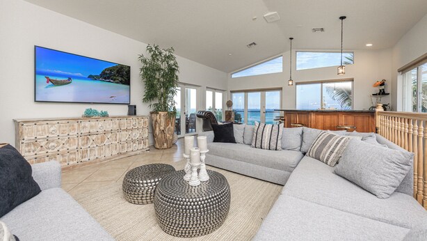 Un salón espacioso con una amplia televisión de pantalla plana, un sofá seccional gris cómodo, un mueble de madera y un gran ventanal con vista al océano.