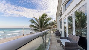 Un balcón con vista a la playa y al océano, amueblado con mobiliario de mimbre y barandilla de cristal.