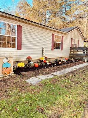 A house with a welcoming sign, a garden with various plants and pumpkins, and a wooden bench.