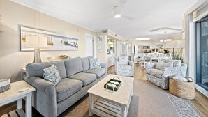 Ein modernes Wohnzimmer mit grauem Sofa, einem passenden Sessel und einem Couchtisch. Im Hintergrund befindet sich eine Küche mit weißen Schränken und einem Essbereich.