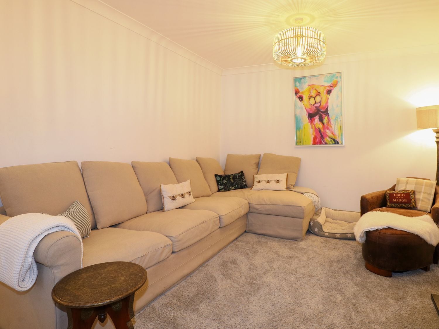 A living room with a beige sectional sofa, a small round wooden table, a brown leather ottoman, and a colorful painting on the wall.