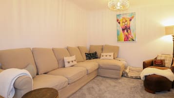 A living room with a beige sectional sofa, a small round wooden table, a brown leather ottoman, and a colorful painting on the wall.