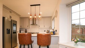 A modern kitchen with stainless steel appliances, a central island with stools, pendant lighting, and a view of a brick wall through a window. - Number 5 (Chichester)