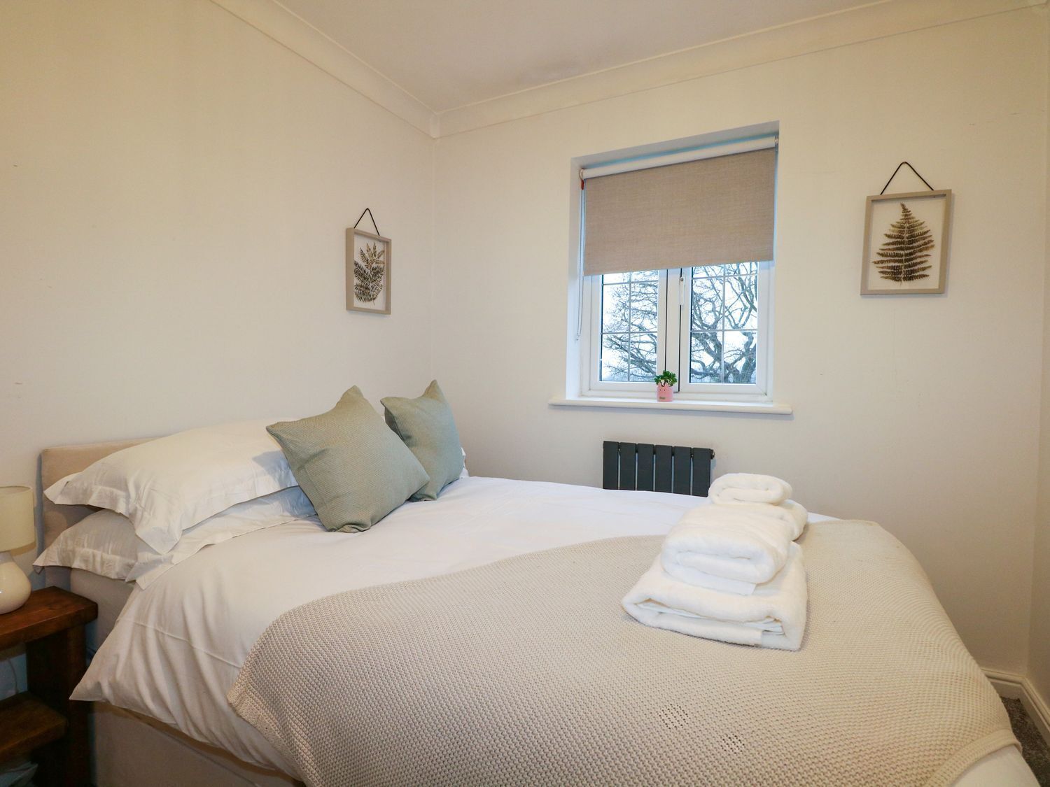 Una cama bien hecha con sábanas blancas y dos almohadas, una lámpara de noche y una ventana con vistas a árboles.