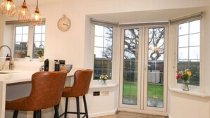A kitchen with a white countertop, two brown bar stools, a clock, and a view of the backyard through glass doors. - Number 5 (Chichester)