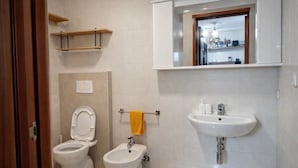 A bathroom with a toilet, bidet, and sink. There are shelves above the toilet and a mirror above the sink. - Hotiday Monopoli Collina (Monopoli)