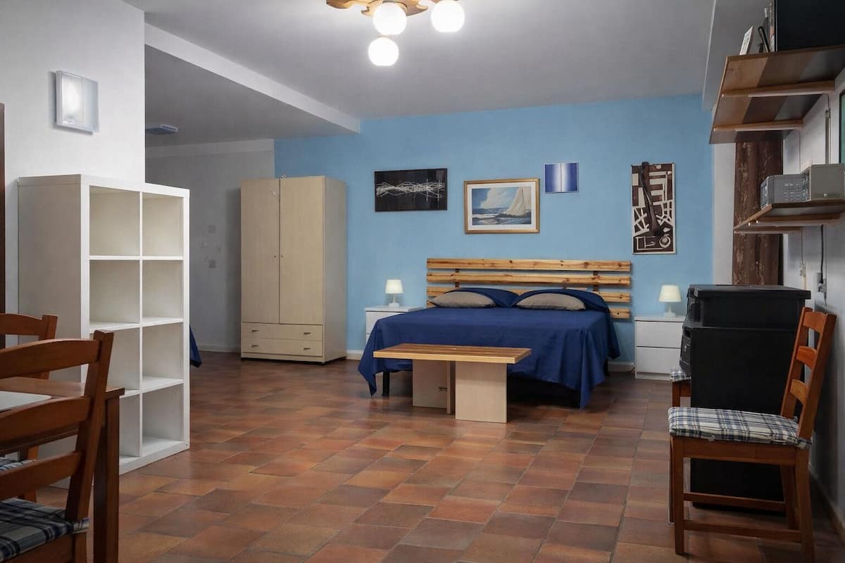 A bedroom with a bed, a desk, a chair, a wardrobe, and a shelf with items.