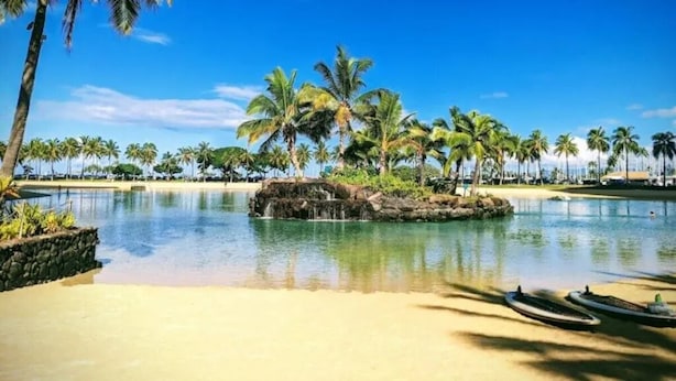 A tropical resort with a clear pool, palm trees, and a rocky island with a waterfall.