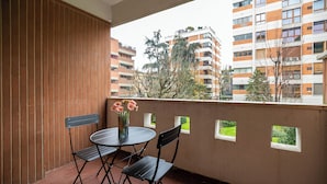 Un balcón con una mesa redonda y dos sillas, un florero con flores, y vistas a edificios de varios pisos.
