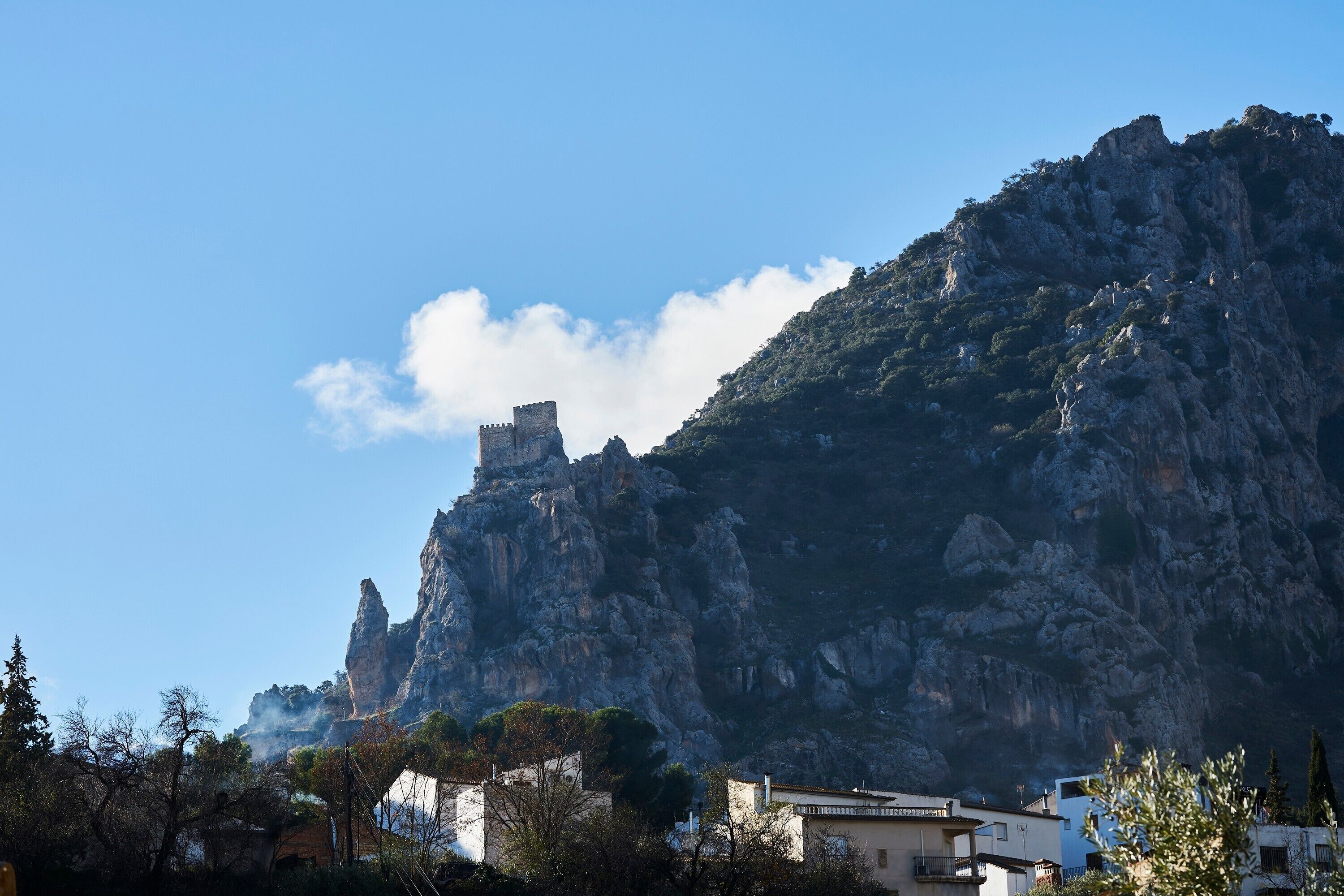 Un paisaje montañoso con una estructura similar a un castillo en un acantilado, rodeado de árboles y edificios.
