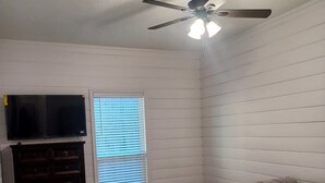 Une chambre à coucher avec un lit, un ventilateur de plafond, une fenêtre avec des stores et un téléviseur posé sur une commode.