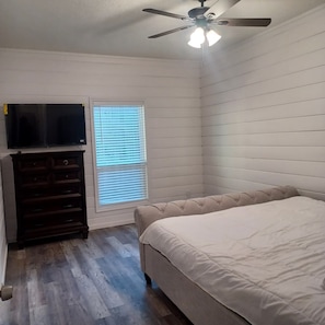 A bedroom with a bed, a ceiling fan, a window with blinds, and a TV on a dresser. - Enjoy the entire private cottage with two comfortable bedrooms, a fully equipped kitchen, spacious living room, full bathroom, and in-unit washer and dryer. Relax with an 85-inch Smart TV, fast Wi-Fi, and comfortable furnishings throughout. (Conroe)