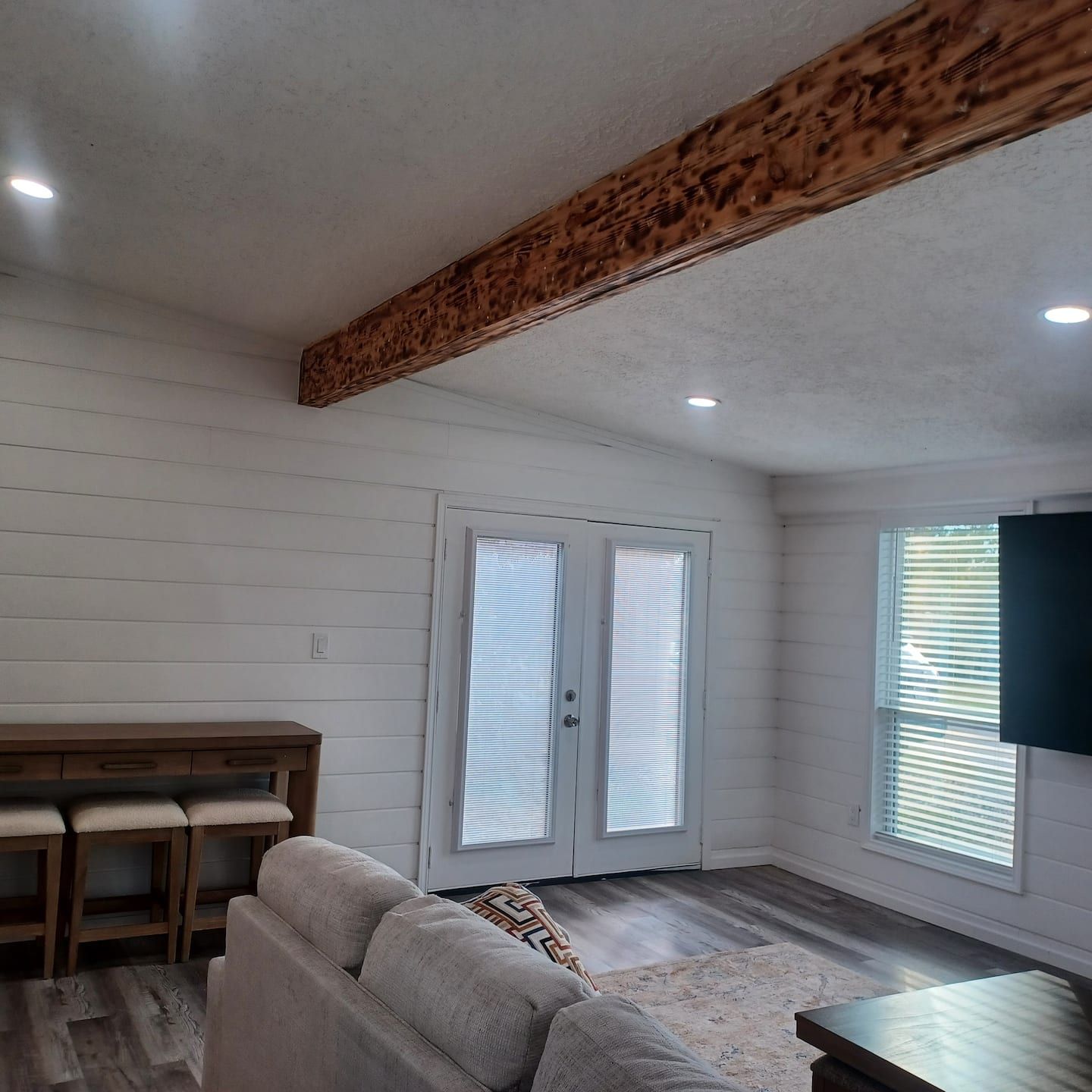 A room with a wooden ceiling beam, a white door with blinds, a sofa, a small table, and a TV mounted on the wall.