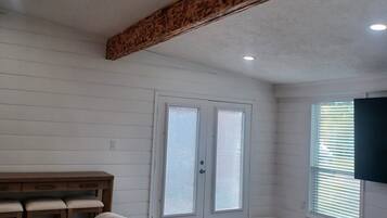 Una habitación con una viga de techo de madera, una puerta blanca con persianas, un sofá, una mesita y un televisor montado en la pared.