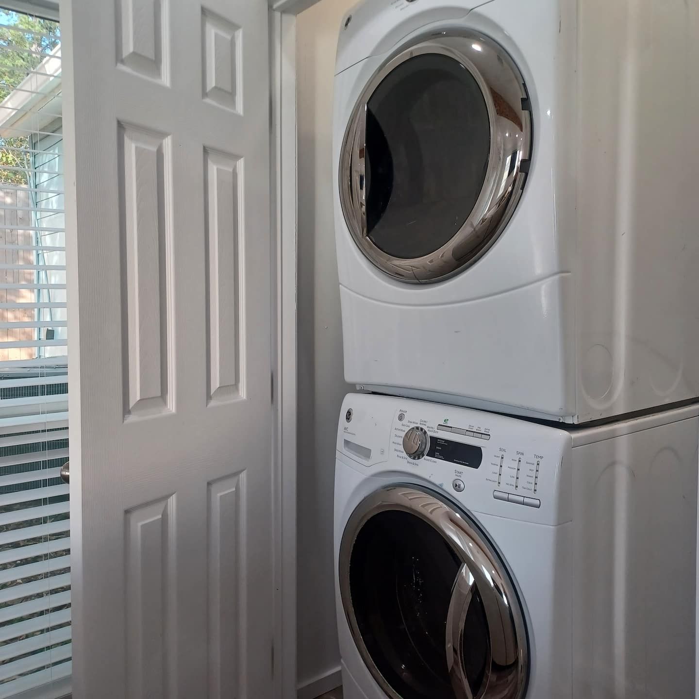 Een gestapelde wasmachine en droger in een ruimte met een witte deur en luiken.