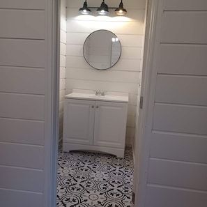 A bathroom with a white vanity, a round mirror, and a patterned floor. - Enjoy the entire private cottage with two comfortable bedrooms, a fully equipped kitchen, spacious living room, full bathroom, and in-unit washer and dryer. Relax with an 85-inch Smart TV, fast Wi-Fi, and comfortable furnishings throughout. (Conroe)