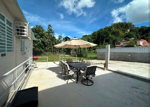 An outdoor patio with a table and chairs, a large umbrella, and a view of a house and trees in the background. - Casa Qodesh #2: Oasis Eco-Gem, Solar & Water (Arecibo)