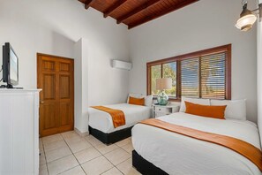 Ein Hotelzimmer mit zwei Betten, einer Holztür, einem Fenster mit Jalousien, einem Nachttisch mit Lampe und einem Fernseher auf einem Schrank.