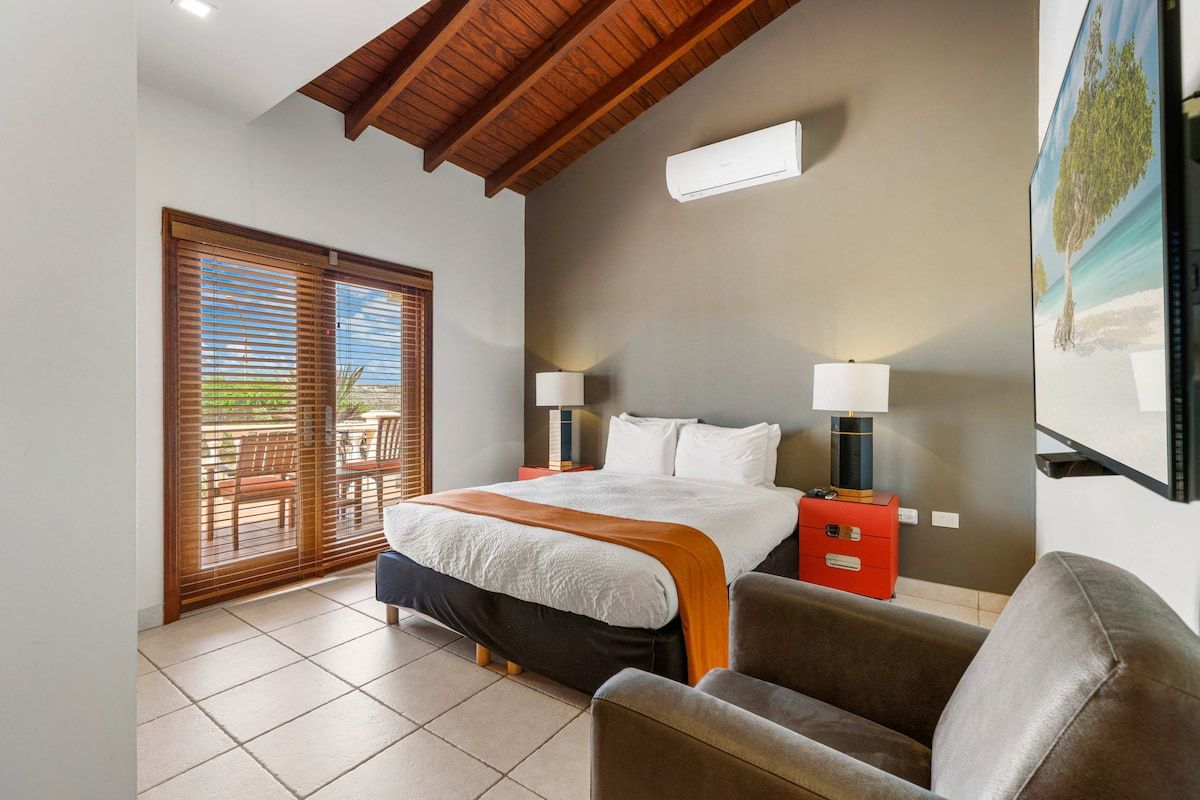 A bedroom with a bed, two bedside lamps, a red nightstand, and a large window with blinds.