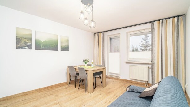 A modern, well-lit room with a wooden floor, a dining table with chairs, a blue sofa, and a window with curtains. - Kraków Huculska Apt — Balcony, Workspace (Kraków)