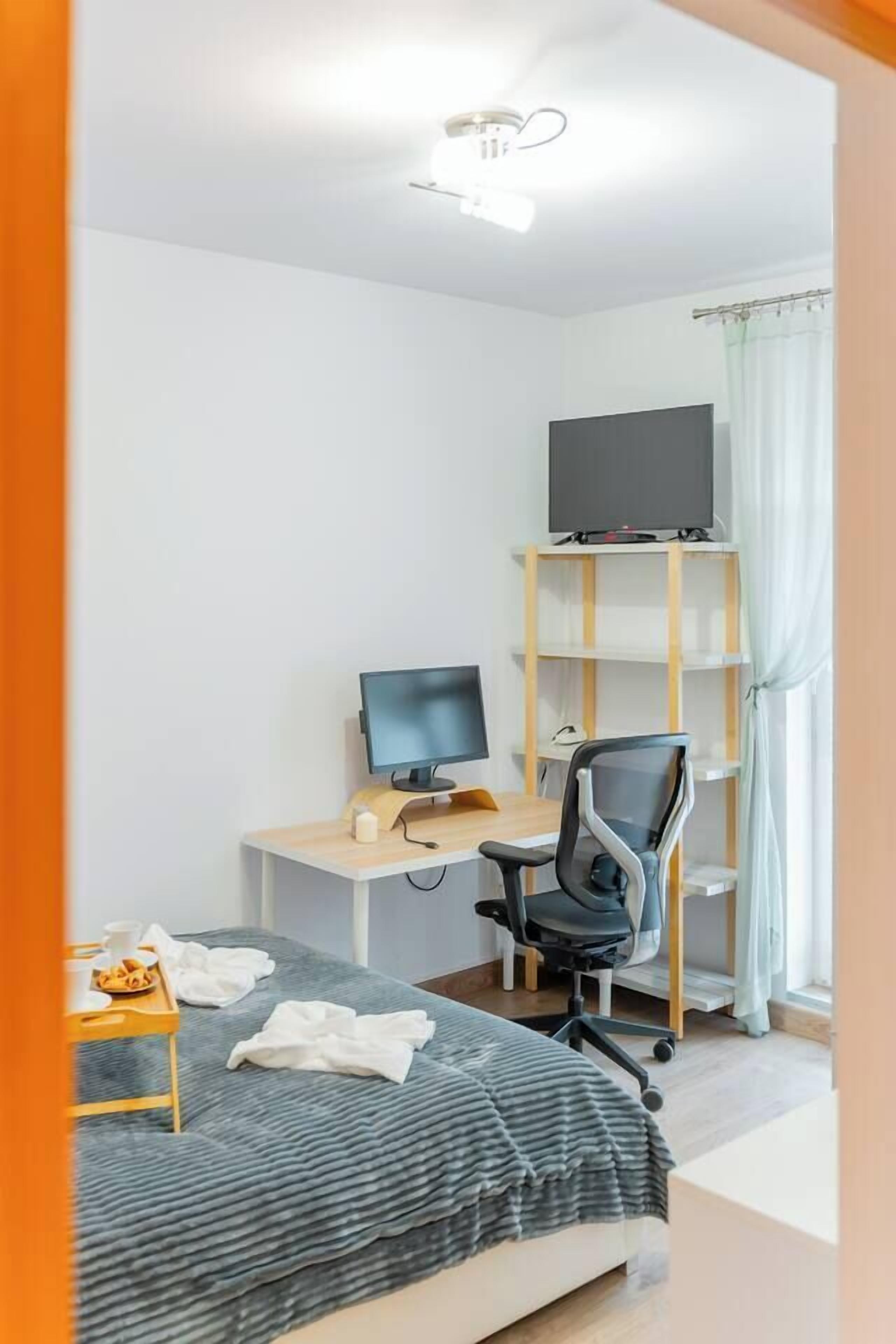 A bedroom with a bed, a desk with a computer, a chair, a TV on a shelf, and a window with curtains.