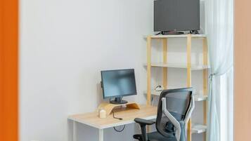 A bedroom with a bed, a desk with a computer, a chair, a TV on a shelf, and a window with curtains.
