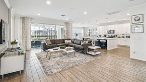 Ein modernes Wohnzimmer mit einer Eckcouch, einem Couchtisch und einem Glastisch. Es gibt eine Küchenzeile mit weißen Schränken und einer Mikrowelle. Das Zimmer verfügt über große Fenster mit Vorhängen.