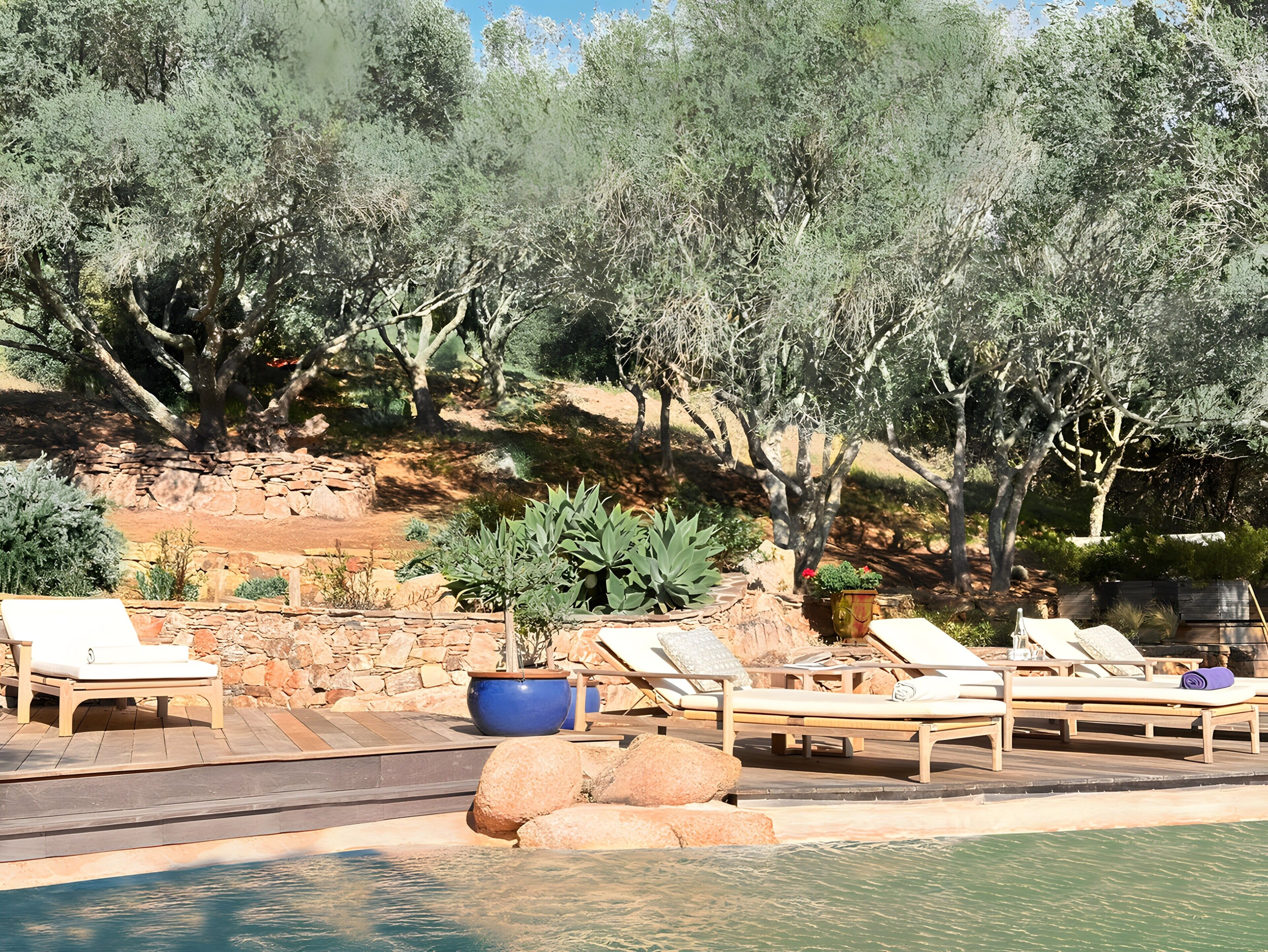 Zona de piscina con sillones, un muro de piedra y un paisaje agreste con árboles.