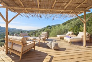 Una terraza de madera con mobiliario exterior, techo de paja y vistas a una ladera panorámica.
