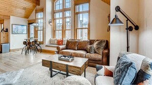 Ein geräumiges Wohnzimmer mit hoher Decke, großen Fenstern, einem Ledersofa, einem Couchtisch und einem Essbereich mit Stühlen.
