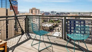 A balcony with two green chairs and a wooden table, overlooking a cityscape with buildings and a clear sky. - Art Lover's Dream: 2BD Luxury 30+ Day, Coors Field (Denver)