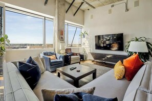 A modern living room with a large TV, comfortable seating, and large windows offering a city view. - Art Lover's Dream: 2BD Luxury 30+ Day, Coors Field (Denver)