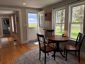 A dining area with a round table and four chairs, a wooden floor, a large window with a view of greenery, and a door leading to another room.