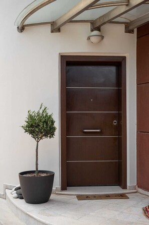 Ein moderner Gebäudeeingang mit einer dunkelbraunen Tür, einem Kübelbaum und einem Metallgeländer.