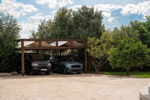Zwei Autos stehen unter einer Holzpergola in einem Garten mit Bäumen und blauem Himmel.